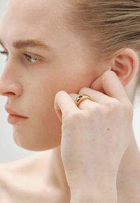 Anillo de oro con un diseño liso que presenta una pequeña gema oscura. Usado en el dedo, exhibido contra un fondo neutro.