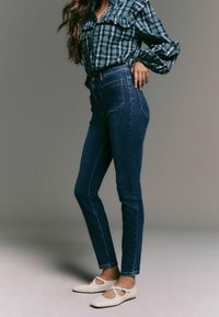 Džíny s vysokým pasem v tmavě modré barvě, s pěti kapsami a přiléhavým střihem, zkombinované se zelenou kostkovanou košilí na knoflíky a bílými balerínkami.