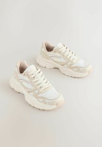 Scarpe da ginnastica bianche e beige con rete e sovrapposizioni sintetiche, punta rotonda, lacci piatti e suola spessa con dettagli testurizzati.