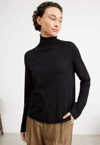Schwarzer Rollkragenpullover aus weichem, strukturiertem Stoff mit langen Ärmeln. Hat einen lockeren Schnitt und einen abgerundeten Saum. Kombiniert mit braunen Hosen.