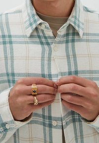 Chemise en flanelle à carreaux crème, bleu sarcelle et beige ; bagues dorées aux doigts, dont une ornée d'une pierre noire et de détails complexes.