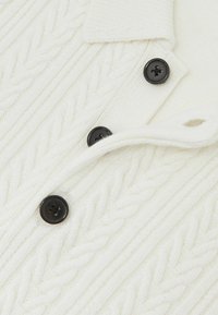 Weißer Strickpullover mit Zopfmuster, einem Kragen und schwarzen Knöpfen. Die Textur ist weich und hat einen gerippten Abschluss.