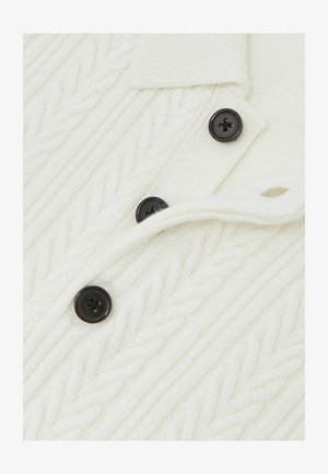Maglione bianco lavorato a maglia con motivo a treccia, colletto e bottoni neri. La texture è morbida con una finitura a costine.