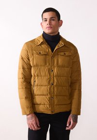 Mustard yellow puffer jacket with quilted texture, two chest pockets, and side zip pockets. Features a button-up front and collar.