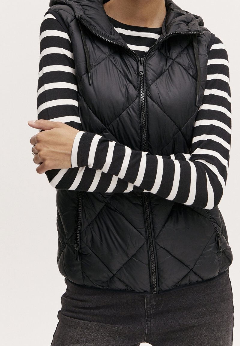 Černá prošívaná péřová vesta s kapucí, předním zipem a fleecovou podšívkou, nošená přes dlouhý rukáv s černobílým pruhovaným vzorem.