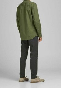 Mies, joka on pukeutunut oliivinvihreään pitkähihaiseen paitaan, tummanharmaisiin housuihin ja beigeihin mokkanahkakenkiin, seisoo selin yksinkertaista harmaata taustaa vasten.