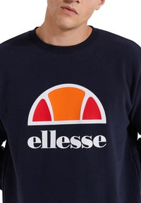 Sudadera azul marino con un logotipo que presenta un semicírculo en naranja y rojo sobre el texto "ellesse" en letras blancas en negrita.