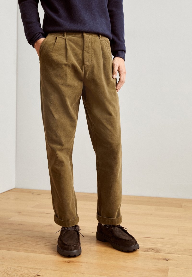 Homme portant un pantalon en velours côtelé marron retroussé aux ourlets, des chaussures à lacets marron foncé et un pull bleu marine, debout sur un sol en bois.