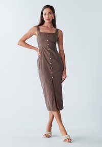 Braunes, knielanges Kleid mit quadratischem Ausschnitt, Knöpfen vorne und seitlichem Schlitz. Das Modell trägt geflochtene Sandalen. Glatte Stofftextur.