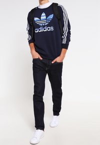 Mörkblå sweatshirt med randiga ärmar och en stor blå och vit Adidas-logotyp, tillsammans med mörka jeans och vita sneakers.