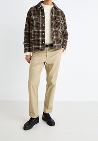 Mężczyzna w brązowej kratkowanej kurtce, białym swetrze, beżowych spodniach, czarnym pasku i czarnych sneakersach stoi z jedną ręką w kieszeni.