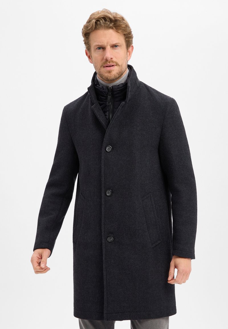 Dunkler Wollmischmantel mit Reverskragen, fünf Knöpfen und zwei seitlichen Taschen. Verfügt über eine schwarz gepolsterte Innenschicht.