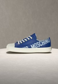 Baskets basses bleues avec lacets et semelle blanches, affichant un grand texte blanc "MOSCHINO" sur le côté.