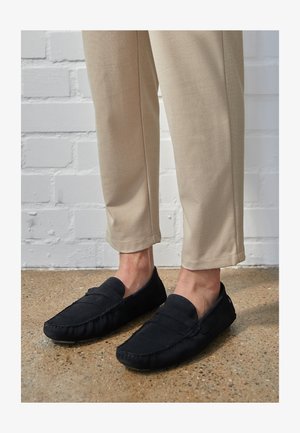 Mocassini neri in suede con finitura testurizzata, caratterizzati da dettagli cuciti e un profilo basso, indossati con pantaloni beige a sigaretta contro un muro bianco.