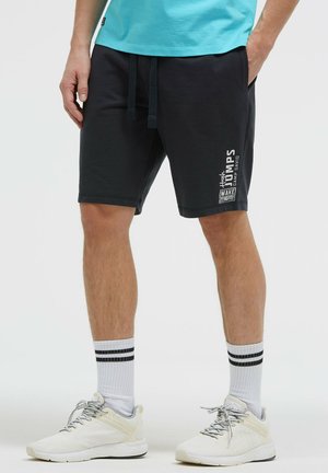 Mann trägt schwarze Freizeithose mit weißem Aufdruck, weiße Sportschuhe und weiße Socken mit zwei schwarzen Streifen, steht mit Händen in den Taschen.