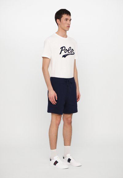 T-shirt branca de algodão com logotipo "Polo" em azul-marinho, combinada com calções de cordão azul-marinho. O modelo usa sapatilhas brancas com detalhes em azul-marinho e meias claras.