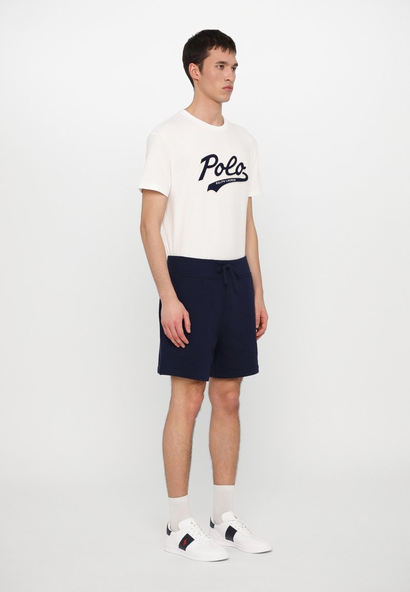 T-shirt en coton blanc avec le logo "Polo" en marine, associé à un short en marine avec cordon de serrage. Le modèle porte des baskets blanches avec des accents en marine et des chaussettes claires.