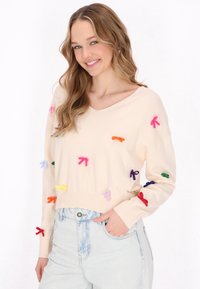 Maglione lavorato a maglia color crema con scollatura a V, decorato con applicazioni di fiocchi multicolori, abbinato a jeans in denim chiaro.