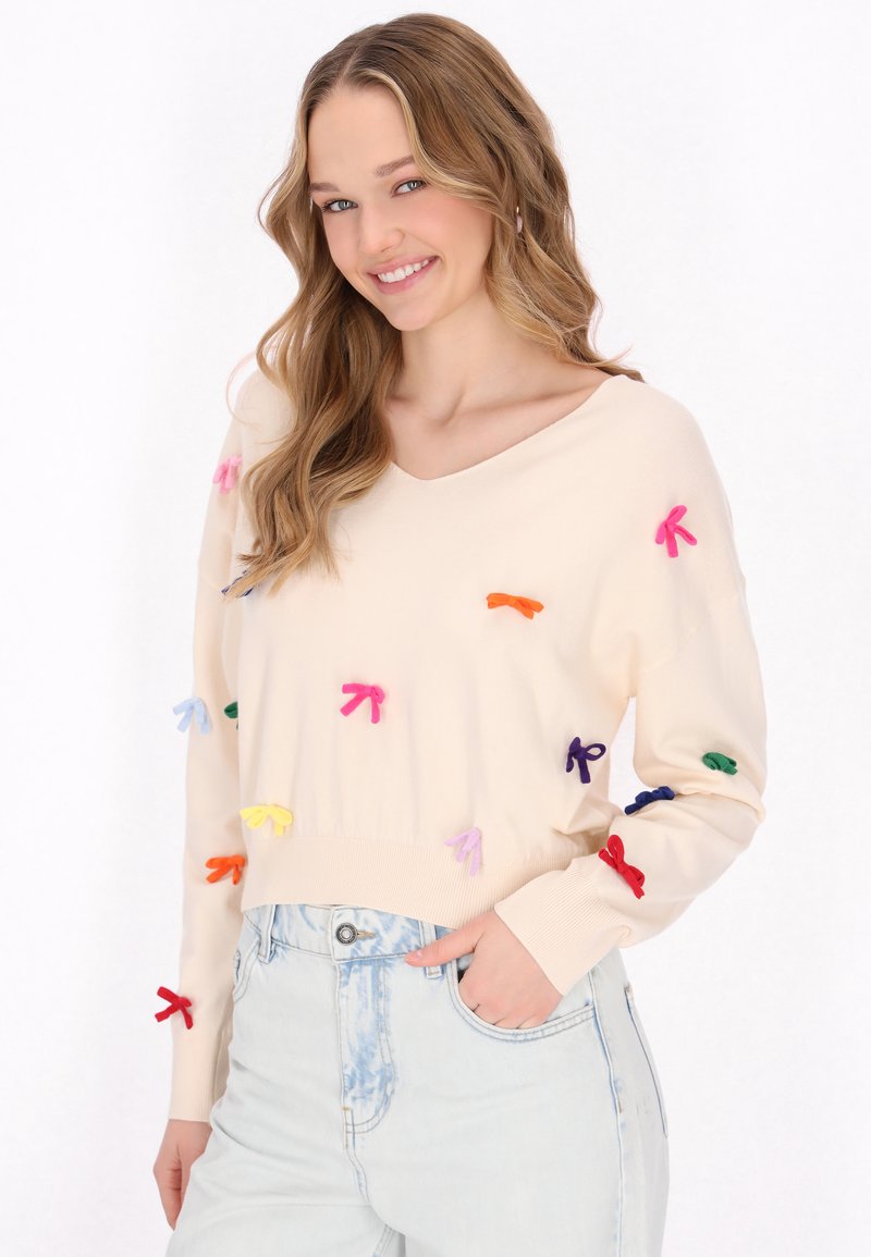Maglione lavorato a maglia color crema con scollatura a V, decorato con applicazioni di fiocchi multicolori, abbinato a jeans in denim chiaro.