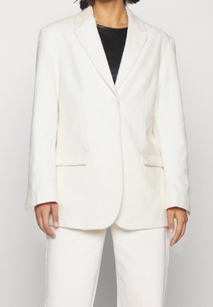 Persona que lleva una blazer blanca de pana sobre una camiseta negra, combinada con pantalones blancos a juego, de pie frente a un fondo liso.