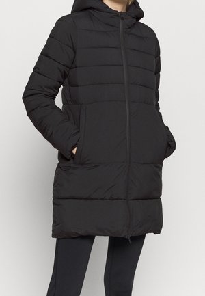 Person trägt einen schwarzen, gesteppte Wintermantel mit Kapuze, Hände in den seitlichen Taschen und schwarze Leggings vor hellem einfarbigem Hintergrund.