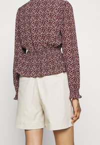 Femme portant un short beige et un blouse à manches longues bordeaux à motifs floraux, montrée de dos avec un petit tatouage sur la main.