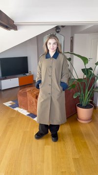 Beige lang kåpe med mørkeblå krage og mansjetter, fem knapper, store lommer, båret over svarte bukser, stående i et rom med lyst tregulv.