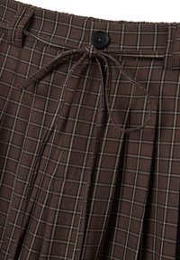 Pantalones de cuadros marrones con cierre de botón, cintura con cordón y diseño plisado. El material parece tener textura con líneas claras contrastantes en el patrón.