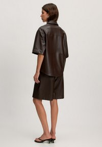 Braune Kunstleder-Übergröße-Hemd mit kurzen Ärmeln, kombiniert mit passenden Shorts. Glatte Textur, lockerer Schnitt und nahtloses Design.