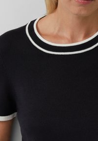 Femme portant un pull noir à manches courtes avec un col en côtes à deux rayures blanches et des bords de manches.