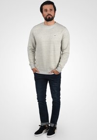 Suéter gris claro con textura sutil, cuello redondo y un pequeño logo. Combinado con jeans de mezclilla oscura y zapatillas negras.