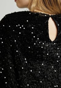 Top noir en sequins avec une apparence texturée et veloutée. Présente une ouverture en forme de clé dans le dos et des sequins circulaires disposés de manière éparse pour plus de détail.