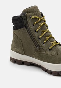 Stivaletto verde in suede con colletto in rete nera testurizzata, lacci gialli, cerniera laterale e suola robusta per una migliore aderenza.