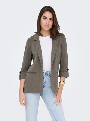 Blazer gris sur mesure en tissu texturé, fermeture à un bouton, manches retroussées et poche avant, porté sur un haut blanc et un jean.