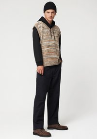 Sweter bez rękawów robiony na drutach z wielokolorowej włóczki, noszony na czarnej bluzie z kapturem. Ciemne spodnie i brązowe buty dopełniają stylizację.