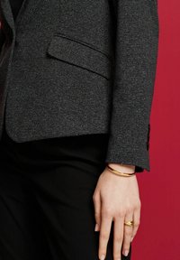 Person som bär en grå strukturerad kavaj och svarta byxor med ett guldarmband och en vriden guldring på vänster hand mot en röd bakgrund.