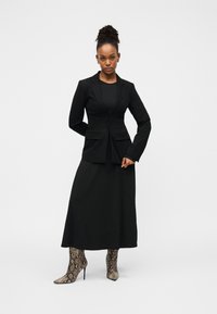 Blazer noir avec revers et deux poches avant, porté sur une longue robe noire. Bottes à talons hauts avec un motif en peau de serpent.