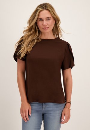 Blonde vrouw met los haar die gouden oorbellen draagt, een bruin kort mouwen topje met pofmouwen en lichtblauwe spijkerbroek.