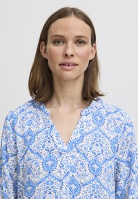 Blusa bianca con stampa floreale blu, caratterizzata da una scollatura a V e una vestibilità morbida. Il tessuto è leggero, con motivi intricati in tutto il capo.
