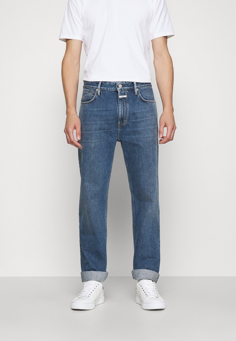 Vaqueros de denim azul con un corte recto, cuatro bolsillos y cierre de botón. Dobladillos recogidos por encima de zapatillas blancas. Camiseta blanca simple.