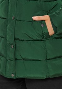 Grön puffjacka med kviltat mönster, med mattsvarta tryckknappar och en sidoficka. Slät textur och rundad krage.