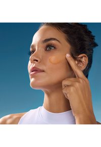 Model nakłada brzoskwiniowy krem na policzek za pomocą opuszka palca, ubrana w jasny top na tle niebieskim.