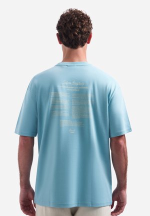 Man met krullend bruin haar, die een lichtblauw oversized T-shirt draagt met witte tekst op de achterkant, staand tegen een effen witte achtergrond.