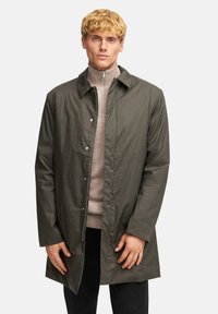 Olivgrön lättviktsjacka med krage, tryckknappar och dragkedja. Bärs över en ljusbeige tröja. Långa ärmar.