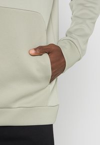 Jasnozielona bluza z teksturowanym wykończeniem, z kieszenią kangurką i ściągaczami na rękawach. Materiał wydaje się miękki i elastyczny.