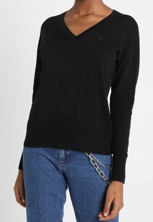 Osoba ubrana w czarny sweter z długim rękawem i dekoltem w serek oraz niebieskie dżinsy z srebrnym łańcuszkiem po prawej stronie.