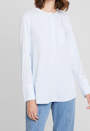 Frau trägt ein hellblau-weiß dünn gestreiftes Langarmhemd mit Tasche und hellblaue Jeans vor neutralem Hintergrund.