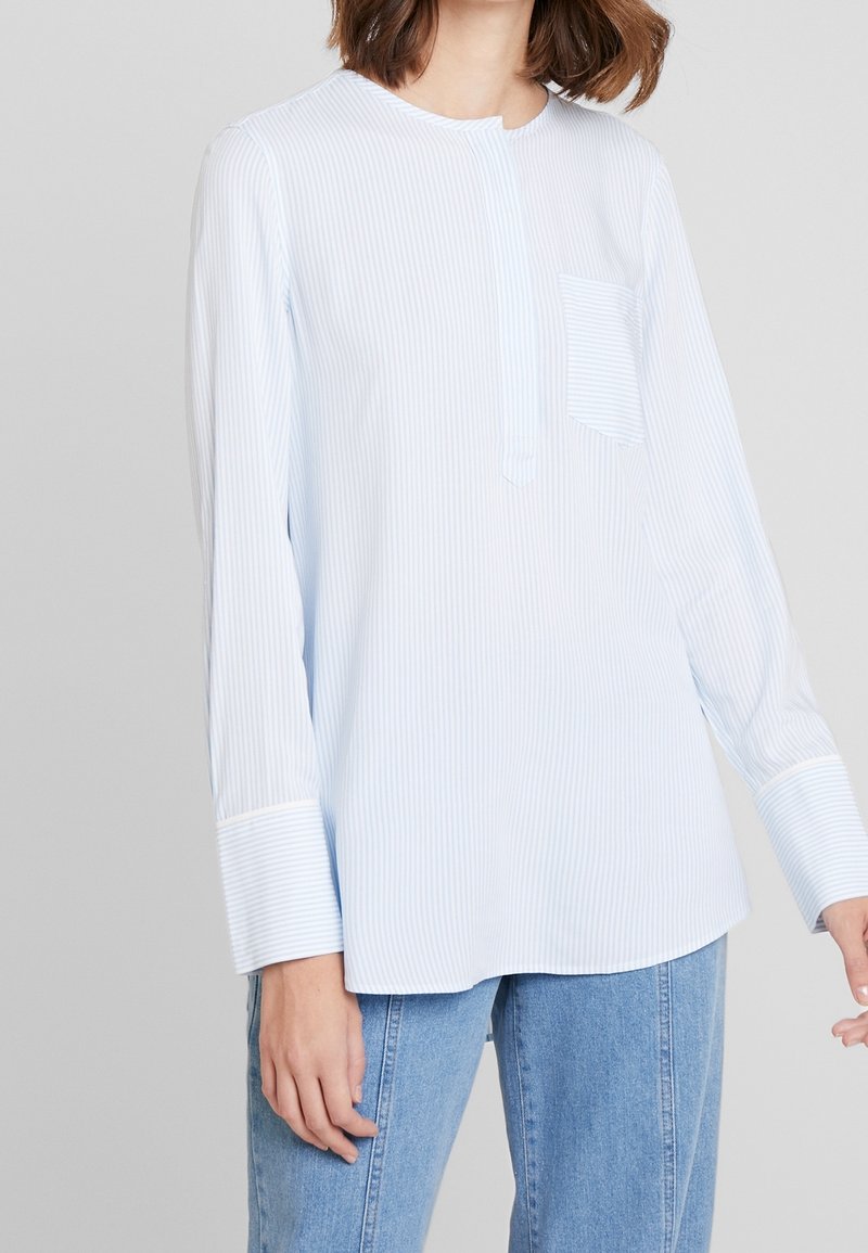 Mujer con una camisa de manga larga de rayas finas en azul claro y blanco, con un bolsillo, y jeans azul claro, sobre un fondo neutro.