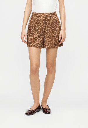 Vrouw draagt bruin korte broek met luipaardprint, wit mouwloos topje en donkerbruine ballerina's, staand tegen een effen lichte achtergrond.