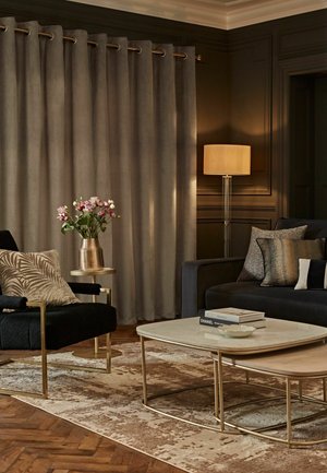 Stue med mørk sofa, sort lænestol med mønstret pude, beige gardiner, gyldigt sidebord med blomster, lagdelte sofaborde og gulvlampe.